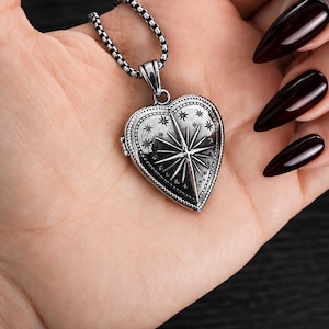 May include: A silver-toned heart-shaped locket pendant featuring an engraved North Star design surrounded by smaller stars. The locket is suspended from a silver-toned box chain necklace, showcasing a detailed metallic finish and a hinged closure.