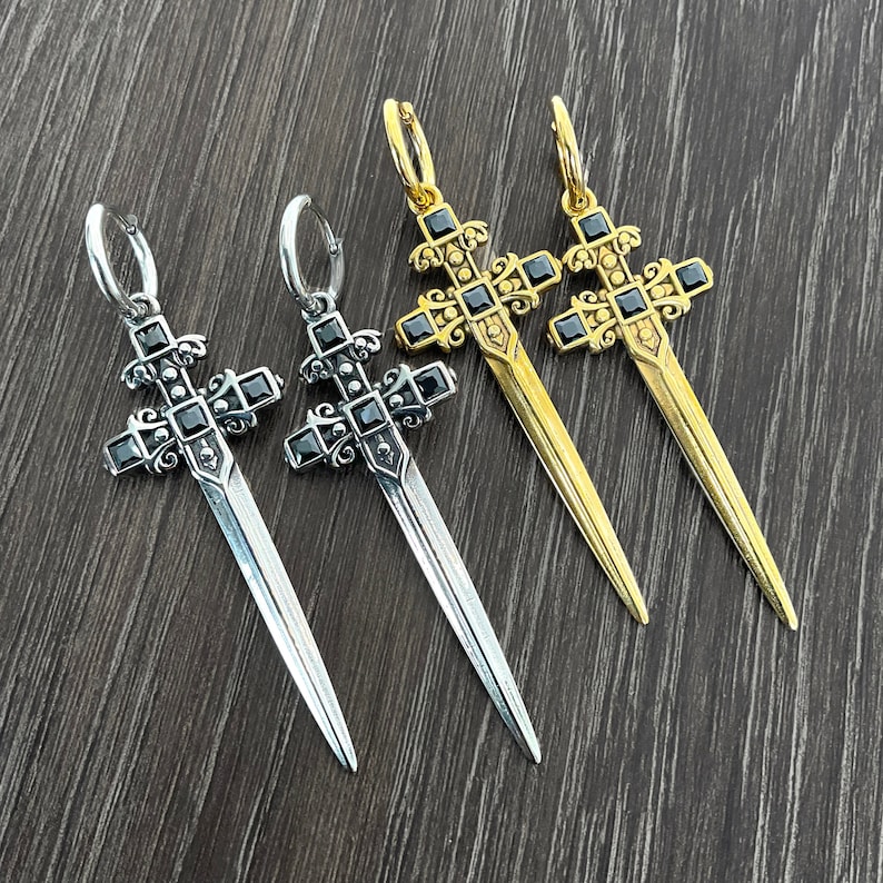 May include: Three silver and gold sword-shaped cross earrings with black gemstones. The earrings are on a wooden surface.