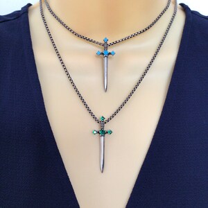 May include: Two silver sword necklaces with turquoise and green gemstones. The necklaces are layered on a model wearing a dark blue top.