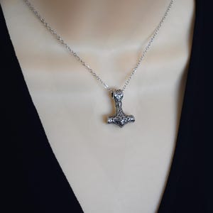 May include: A silver-colored pendant necklace featuring a detailed Thor's hammer design. The hammer pendant is suspended from a delicate chain. The necklace is displayed against a dark background.