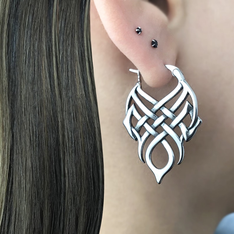 May include: Silver hoop earrings with a detailed Celtic knot design. The earrings are in the shape of a teardrop and have a smooth, polished finish.
