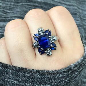 May include: A silver ring featuring a large blue gemstone surrounded by four skull designs. The ring is set with smaller blue gemstones around the main stone.