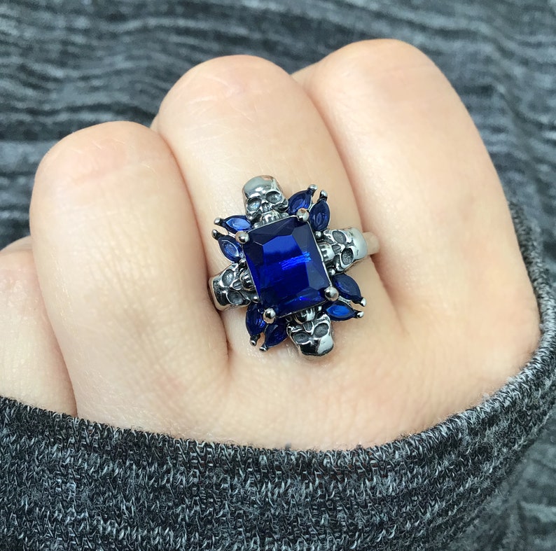 May include: A silver ring featuring a large blue gemstone surrounded by four skull designs. The ring is set with smaller blue gemstones around the main stone.