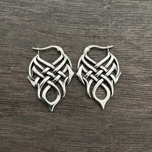 May include: A pair of silver hoop earrings with a detailed, intricate knot design. The earrings are made of metal and have a polished finish.