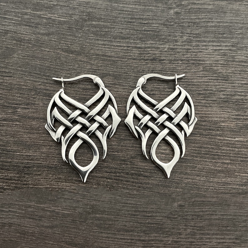 May include: A pair of silver hoop earrings with a detailed, intricate knot design. The earrings are made of metal and have a polished finish.