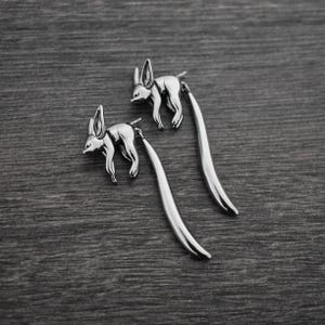 May include: A pair of silver dangle earrings featuring a stylized fennec fox design. The earrings have a long, thin, curved drop.
