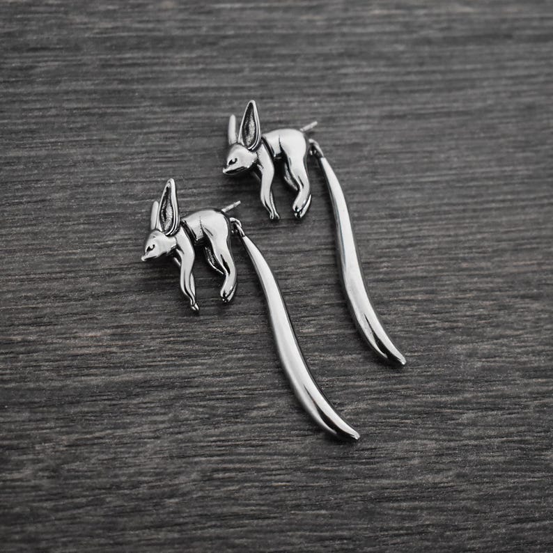 May include: A pair of silver dangle earrings featuring a stylized fennec fox design. The earrings have a long, thin, curved drop.