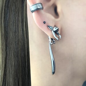 May include: Silver dangle earrings featuring a small fox design with a long tail. The fox is facing forward with its head tilted up. The earrings are a unique and stylish accessory.