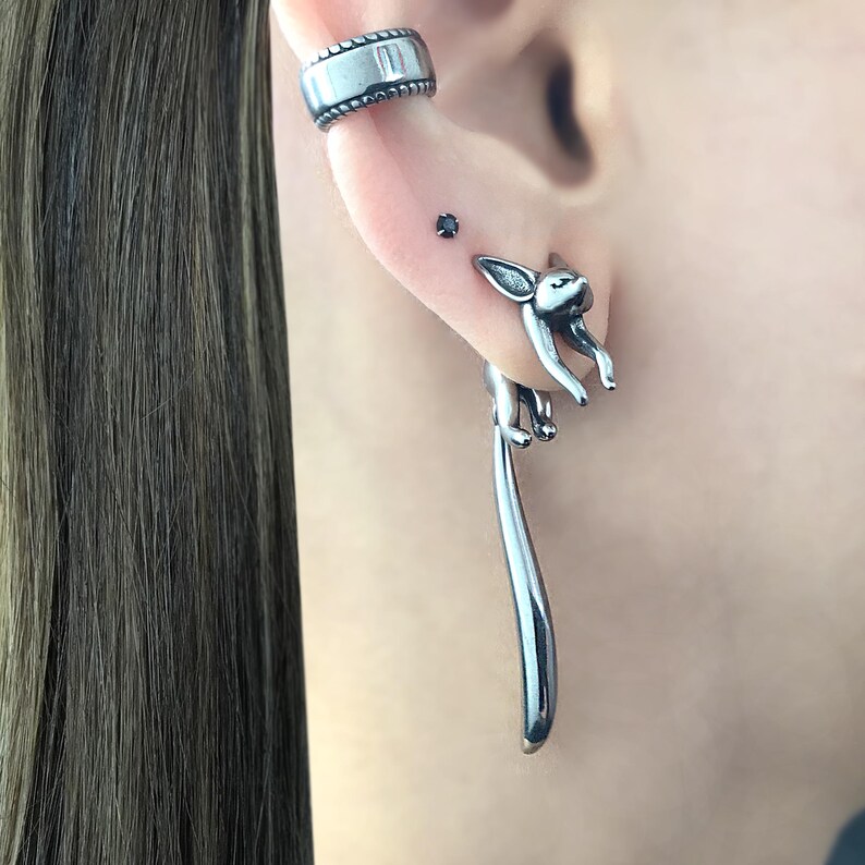 May include: Silver dangle earrings featuring a small fox design with a long tail. The fox is facing forward with its head tilted up. The earrings are a unique and stylish accessory.