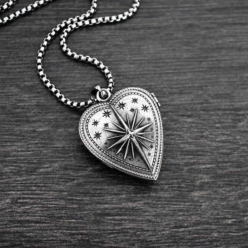 May include: A silver-toned heart-shaped locket pendant suspended from a matching box chain. The locket features an embossed North Star design surrounded by small engraved stars, with a beaded border detail along the outer edge of the heart.