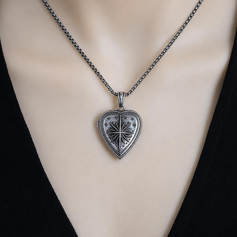 May include: A silver-coloured heart-shaped locket pendant featuring an engraved starburst design surrounded by small stars. The locket hangs from a matching silver-coloured box chain necklace, showcasing an oxidised finish with intricate detailing.