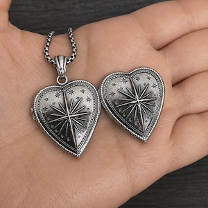 May include: A silver-toned heart-shaped locket pendant featuring an embossed North Star design surrounded by small stars and a beaded border. The locket is attached to a silver-toned chain, and a matching heart-shaped piece is placed beside it.