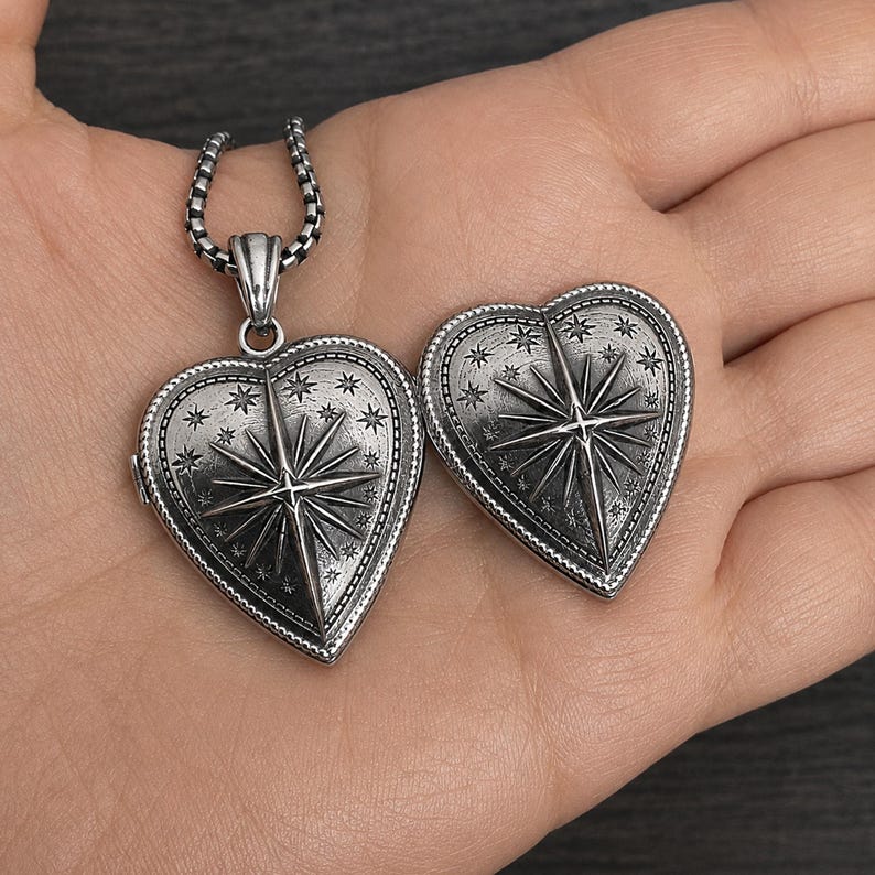 May include: A silver-toned heart-shaped locket pendant featuring an embossed North Star design surrounded by small stars and a beaded border. The locket is attached to a silver-toned chain, and a matching heart-shaped piece is placed beside it.