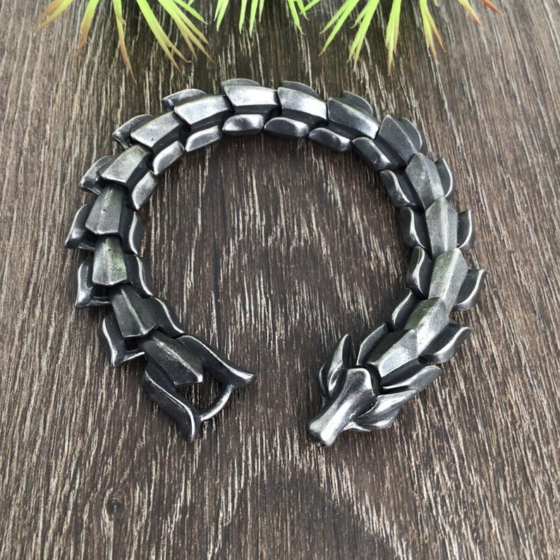 Puede incluir: Una pulsera plateada con un dise&ntilde;o de drag&oacute;n. La pulsera est&aacute; hecha de metal y tiene una superficie texturizada.