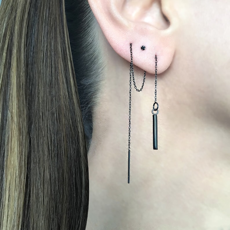 May include: Black chain earrings with a small bar pendant hanging from one chain. The earrings are worn in the earlobe and helix piercing.