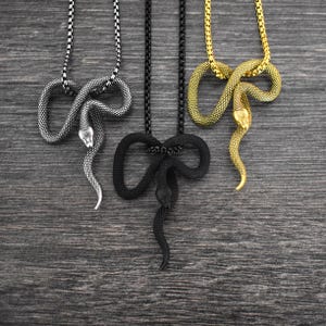 May include: Three snake pendant necklaces in silver, black, and gold. Each necklace features a detailed snake pendant with a matching chain. The pendants are intricately designed, showcasing the snake's form. The necklaces are displayed on a dark wood-grain surface.