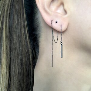 May include: Black chain earrings with two dangling bar charms. The earrings are worn in the left ear.