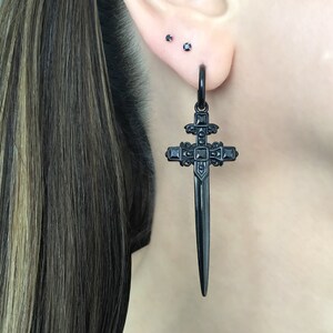 May include: Black sword-shaped cross earring with black cubic zirconia accents. The earring is hanging from a person's earlobe.