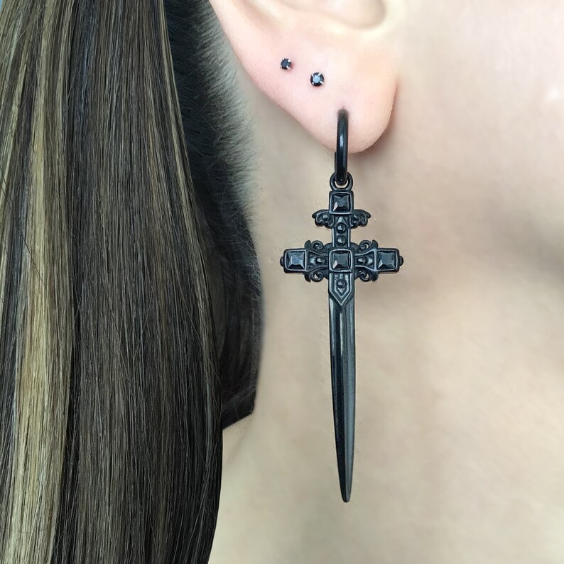 May include: Black sword-shaped cross earring with black cubic zirconia accents. The earring is hanging from a person's earlobe.