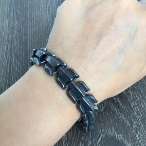 Puede incluir: Un brazalete de metal negro con un dise&ntilde;o &uacute;nico entrelazado. El brazalete est&aacute; hecho de una serie de eslabones conectados que forman un patr&oacute;n geom&eacute;trico.