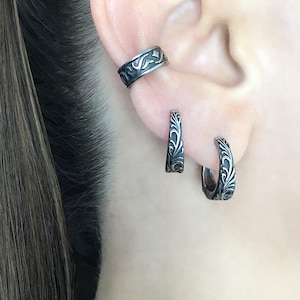 May include: Three silver hoop earrings with an intricate floral design. The earrings are shown on a person's ear.
