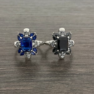 May include: Two silver rings with skull details. One ring has a blue gemstone and the other has a black gemstone.