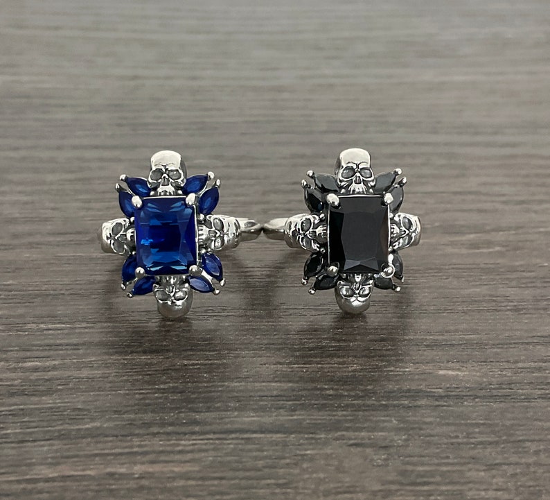 May include: Two silver rings with skull details. One ring has a blue gemstone and the other has a black gemstone.