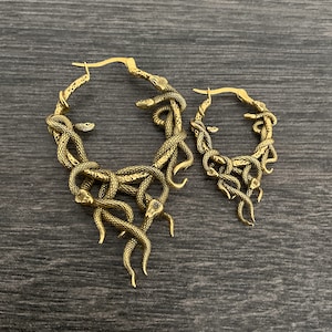 May include: A pair of gold hoop earrings featuring a snake design. The larger earring has multiple snakes intertwined, while the smaller earring has a simpler design with fewer snakes.