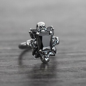 May include: A silver ring featuring a black gemstone surrounded by five skull designs.
