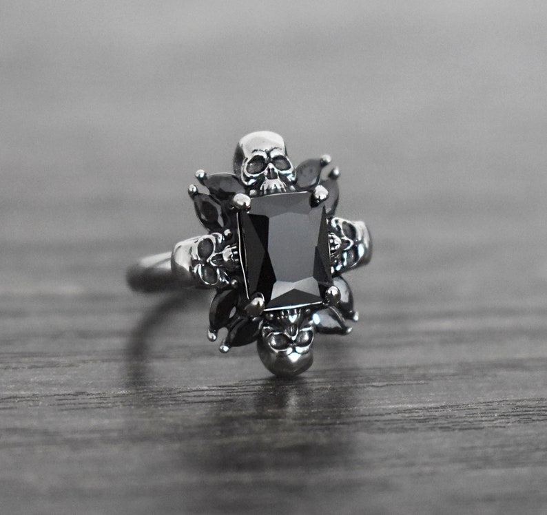 May include: A silver ring featuring a black gemstone surrounded by five skull designs.