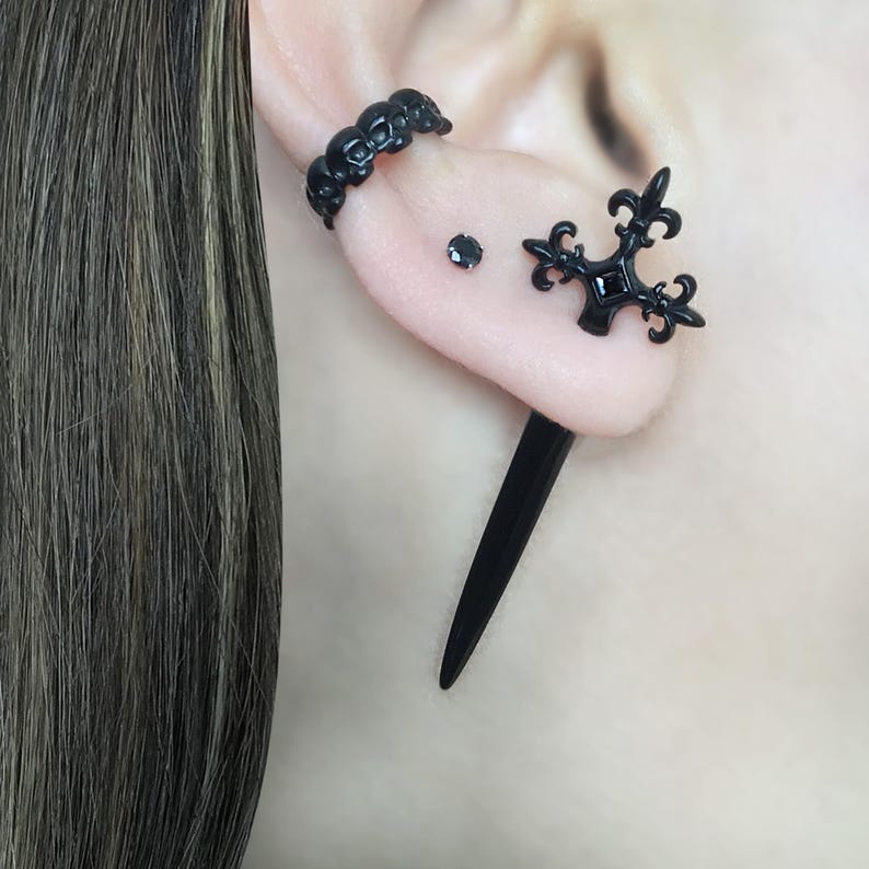 Pu&ograve; includere: Anello nero a forma di teschio, orecchino a croce nero e orecchino a punta nero. Tutti e tre sono indossati nell'orecchio di una persona con i capelli neri.