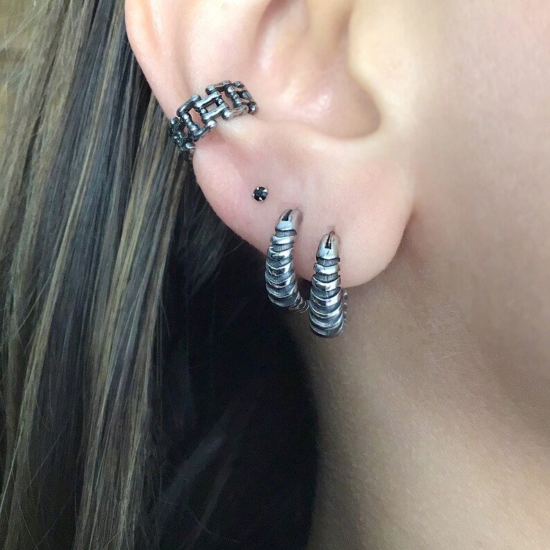May include: A person's ear with three silver earrings. The top earring is a chain link design, the middle earring is a small black stud, and the bottom two earrings are silver hoops with a textured design.