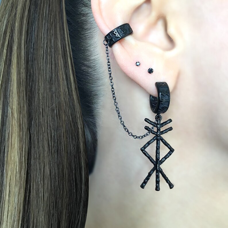 May include: Black ear cuff and earring set. The ear cuff wraps around the upper ear and connects to a hoop earring with a chain. The earring features a stylized symbol. The jewelry is made of black metal.
