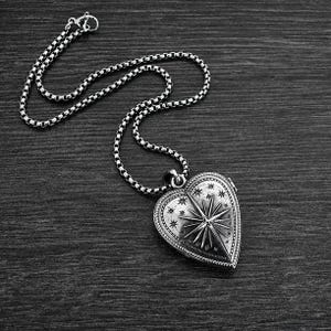 May include: A silver-toned heart-shaped locket pendant suspended from a matching box chain. The locket features an engraved celestial design with a prominent central starburst surrounded by smaller stars and a beaded border on the outer edge.
