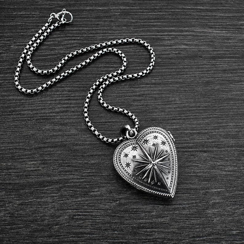 May include: A silver-toned heart-shaped locket pendant suspended from a matching box chain. The locket features an engraved celestial design with a prominent central starburst surrounded by smaller stars and a beaded border on the outer edge.