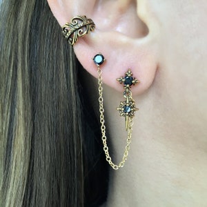 May include: Gold-toned ear cuff and earrings set. The ear cuff features an ornate design with small embellishments. One earring is a stud with a black gem, connected by a chain to a dangling earring with a starburst design and a black gem.