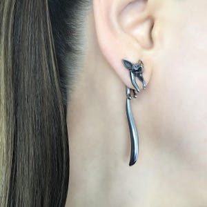 May include: A pair of silver earrings featuring a small, detailed sculpture of a cat with its tail extending down as a long, thin rod.