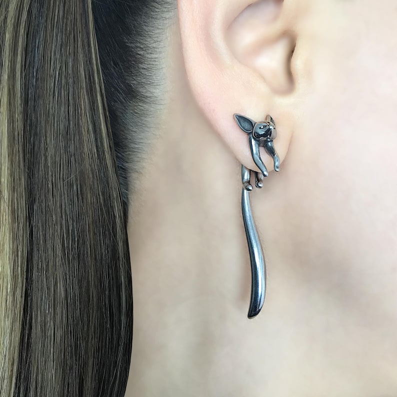 May include: A pair of silver earrings featuring a small, detailed sculpture of a cat with its tail extending down as a long, thin rod.