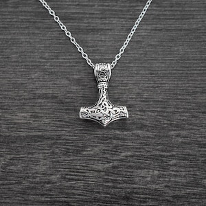 May include: A silver-colored pendant necklace featuring a detailed Thor's hammer design. The hammer pendant is intricately carved with swirling patterns and is suspended from a delicate chain. The necklace is displayed against a dark wood grain background.