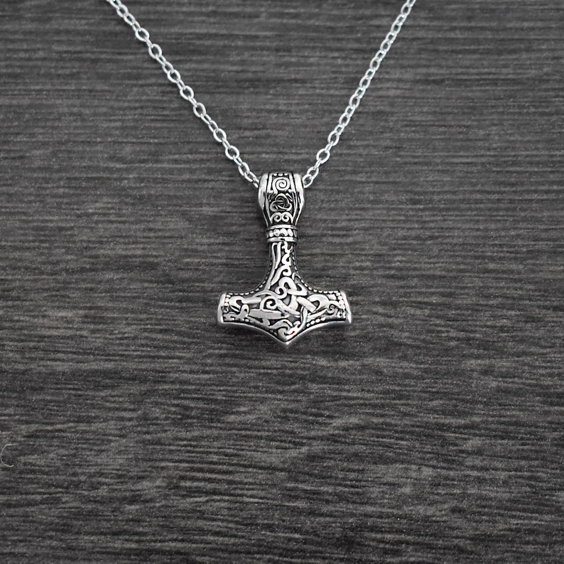 May include: A silver-colored pendant necklace featuring a detailed Thor's hammer design. The hammer pendant is intricately carved with swirling patterns and is suspended from a delicate chain. The necklace is displayed against a dark wood grain background.