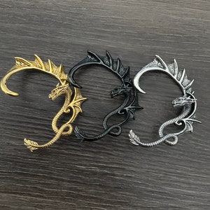 Op de afbeelding: Drie draken oormanchetten in goud, zwart en zilver. Elke manchet heeft een gedetailleerd drakenontwerp met vleugels en een gekrulde staart.