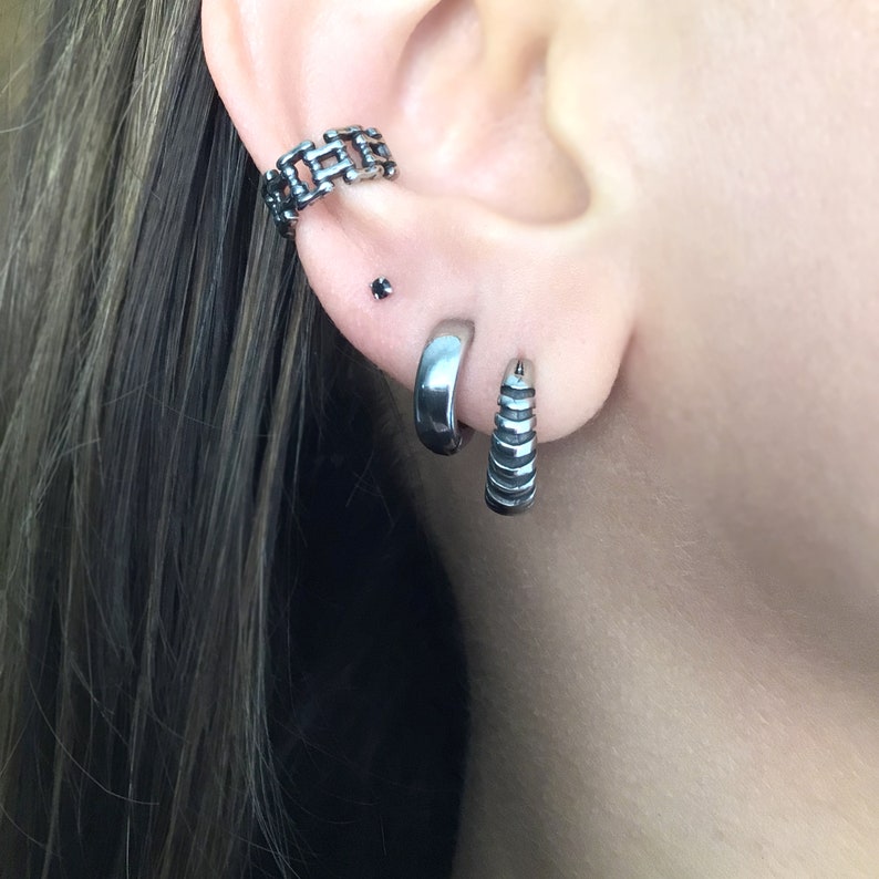 May include: A person's ear with three silver earrings. The top earring is a chain link design, the middle earring is a small stud, and the bottom earring is a hoop with a textured design.