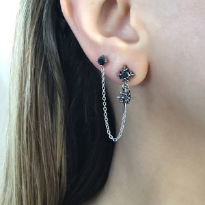 May include: A pair of silver earrings with black gemstones. One earring is a stud, and the other features a starburst design with a chain connecting to a second stud. The earrings are worn on an ear.