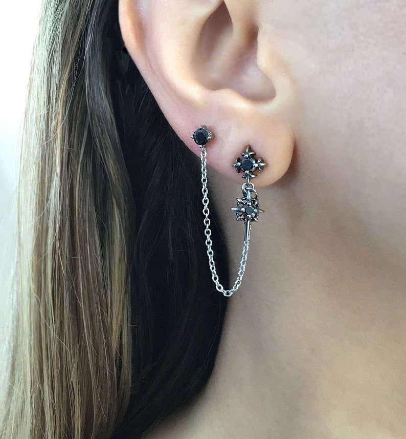 May include: A pair of silver earrings with black gemstones. One earring is a stud, and the other features a starburst design with a chain connecting to a second stud. The earrings are worn on an ear.