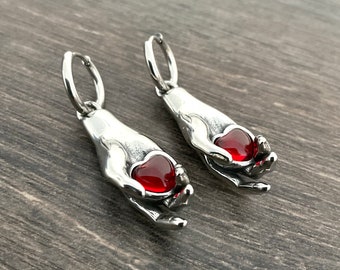 May include: A pair of silver hoop earrings featuring a hand holding a red heart charm. The earrings are made of metal and have a polished finish.