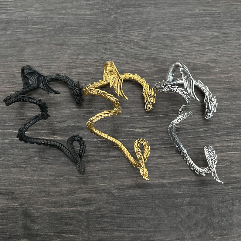 Puede incluir: Tres pendientes de oreja en forma de drag&oacute;n en negro, dorado y plateado. Los pendientes est&aacute;n enrollados y tienen alas.