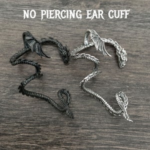 Puede incluir: Dos pendientes de drag&oacute;n, uno negro y otro plateado, se muestran sobre una superficie de madera. Los pendientes est&aacute;n dise&ntilde;ados para envolverse alrededor de la oreja sin perforar. El texto "NO PIERCING EAR CUFF" se muestra sobre los pendientes.