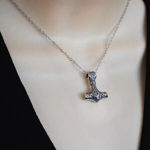 May include: A silver-colored pendant necklace featuring a detailed Thor's hammer design. The pendant hangs from a delicate chain. The hammer has intricate patterns and is a symbol of Norse mythology.