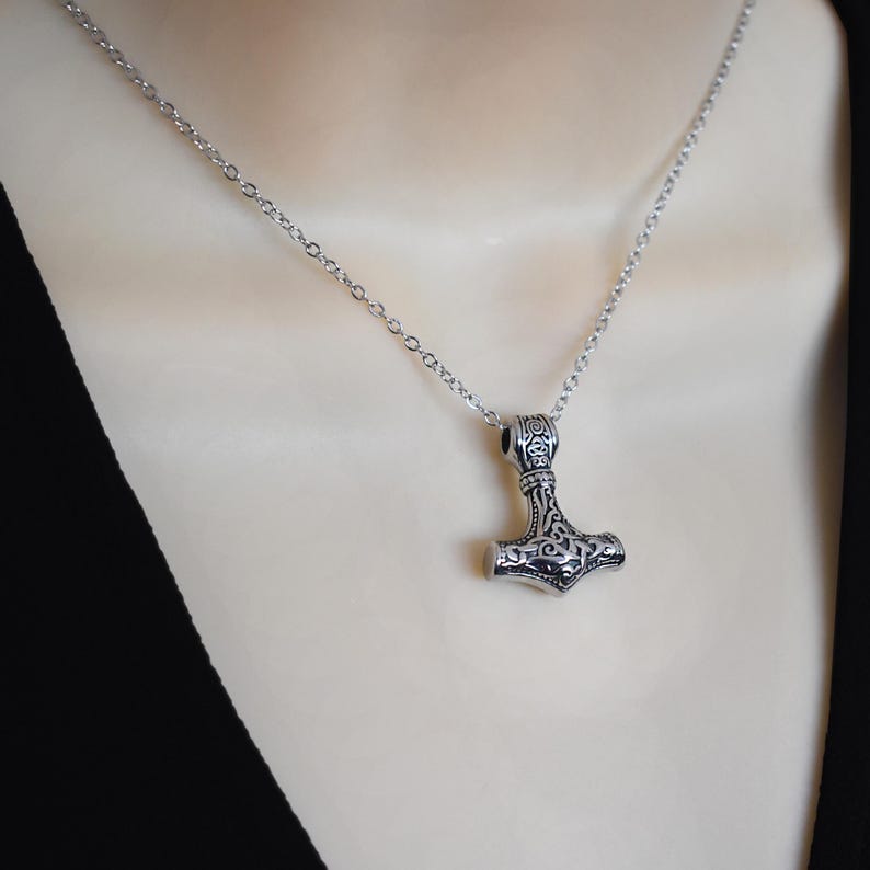 May include: A silver-colored pendant necklace featuring a detailed Thor's hammer design. The pendant hangs from a delicate chain. The hammer has intricate patterns and is a symbol of Norse mythology.