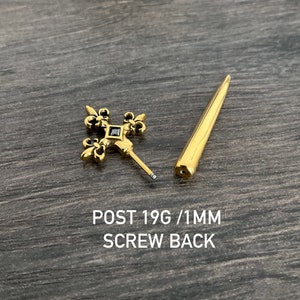 Pu&ograve; includere: Gioiello per il corpo color oro con una croce con dettagli a giglio e una gemma nera al centro. Il gioiello &egrave; fornito con un perno a vite. POST 19G / 1MM SCREW BACK.